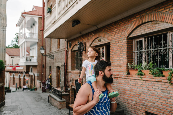 Young father walking with little daughter outdoors, on streets in city of Tbilisi in the capital of Georgia on sunny spring day. Dad and girl travel on excursion in old town. - Fotó, kép