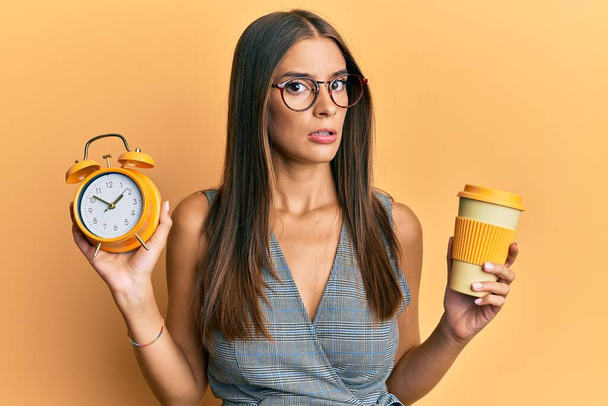 Genç İspanyol kadın çalar saati tutuyor ve bir fincan kahve içiyor habersiz ve kafası karışık bir ifade. şüphe kavramı.  - Fotoğraf, Görsel