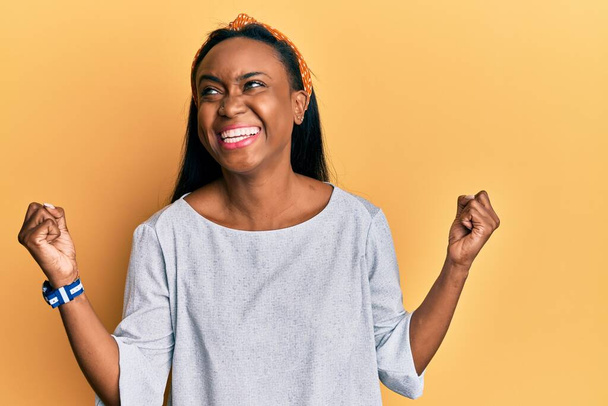 Fiatal afrikai nő visel alkalmi ruhák sárga háttér nagyon boldog és izgatott csinál győztes gesztus felemelt karokkal, mosolyog és sikoltozik a sikerért. ünnepség koncepciója.  - Fotó, kép