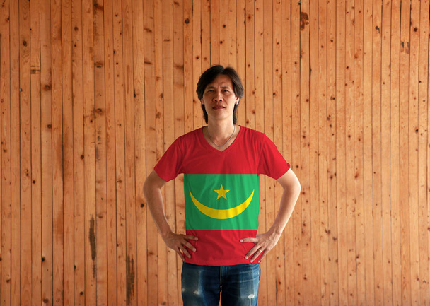 Man wearing Mauritania flag color shirt and standing with akimbo on the wooden wall background, two red stripes flanking a green field with a golden crescent and star.  - Photo, Image