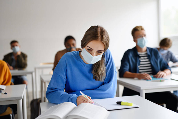 Young students with face masks at desks at college or university, coronavirus concept. - Фото, изображение