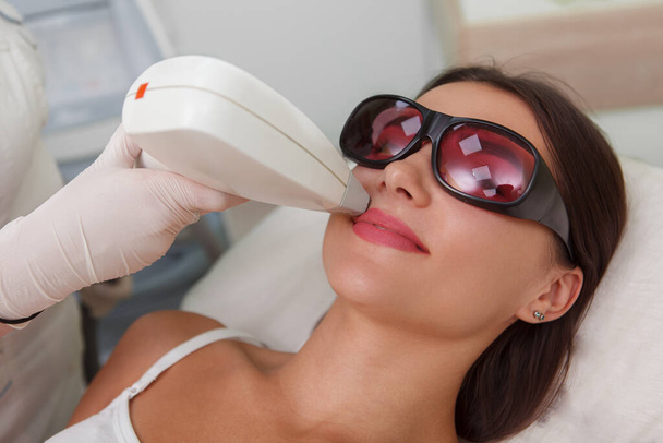 Close up of a woman wearing protective eyeglasses, getting upper lip hair laser remover by cosmetologist - Photo, Image