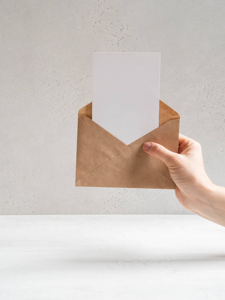 Mão feminina segure o envelope kraft aberto e cartão postal no fundo cinza, vista frontal. Espaço de cópia - Foto, Imagem