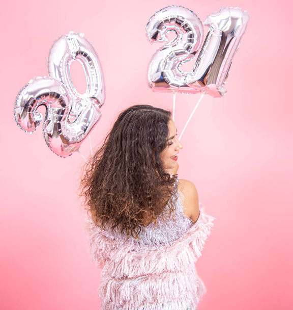 Brunette girl with curly hair in festive clothes from the back posing on a pink background with silver New Year 2021 balls in her hands close up - Photo, Image