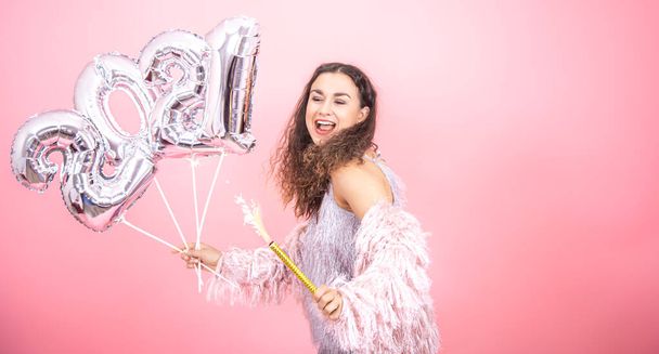 Affascinante giovane ragazza bruna con i capelli ricci festivamente vestita sorridente e tenendo una candela fuochi d'artificio in mano e palloncini d'argento dai numeri 2021 su uno sfondo rosa - Foto, immagini