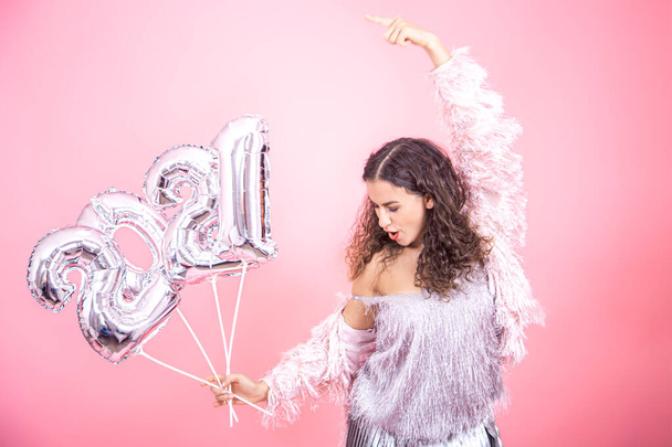 Partygirl Brünette mit lockigem Haar tanzt festlich gekleidet auf rosa Hintergrund mit warmem Licht und silbernen Luftballons in den Händen aus den Nummern 2021 - Foto, Bild