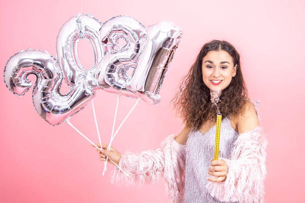 Suloinen juhlallisesti pukeutunut brunette tyttö, jolla on kiharat hiukset vaaleanpunaisella studiotaustalla, katsoo sammutettua ilotulituskynttilää, jossa on hopeailmapalloja numeroista 2021 - Valokuva, kuva