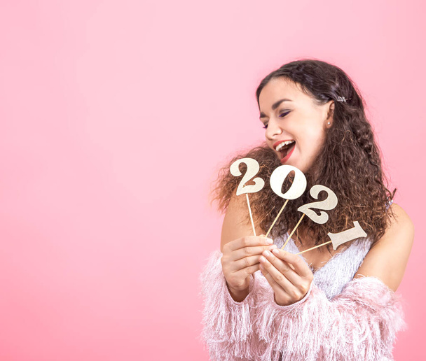Número de madera 2021 en las manos de una hermosa chica alegre en un fondo de estudio rosa, feliz concepto de año nuevo - Foto, Imagen