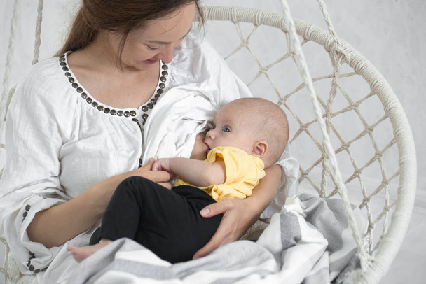 母親は生まれたばかりの赤ん坊を明るい背景の白いハンモックの椅子で母乳で育てます。母乳育児の概念を閉じます. - 写真・画像
