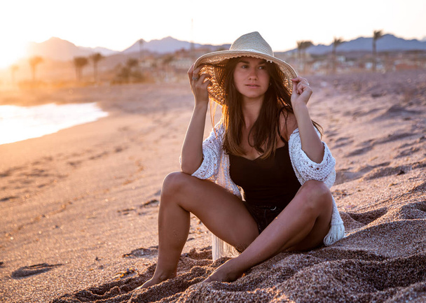 Piękna boho modelka w kostiumie kąpielowym z białą peleryną z kapeluszem na głowie pozuje na plaży o zachodzie słońca - Zdjęcie, obraz