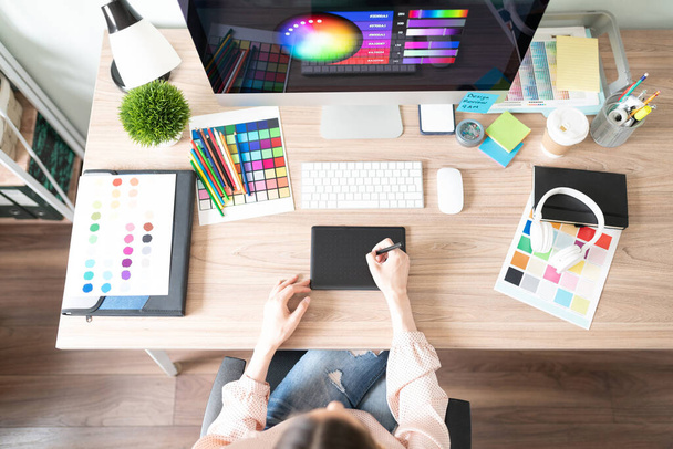 Vista superior de una mujer usando una tableta gráfica y haciendo algún trabajo de diseño en su espacio de trabajo con muchas paletas de colores y muestras - Foto, Imagen