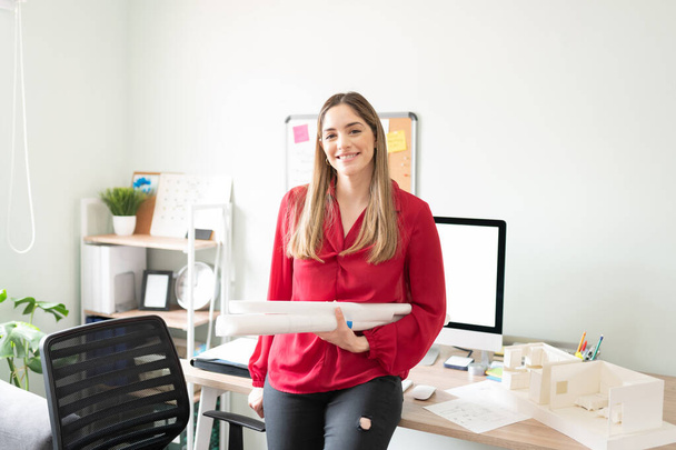 Красивая женщина-архитектор держит несколько свернутых планов, сидя на столе в офисе и улыбаясь - Фото, изображение