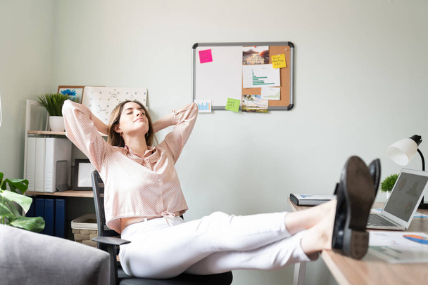 Ganzkörperansicht einer Frau, die lässig und entspannt in ihrem Büro gekleidet ist, die Füße auf den Schreibtisch legt und sich auf ihren Stuhl zurücklehnt - Foto, Bild