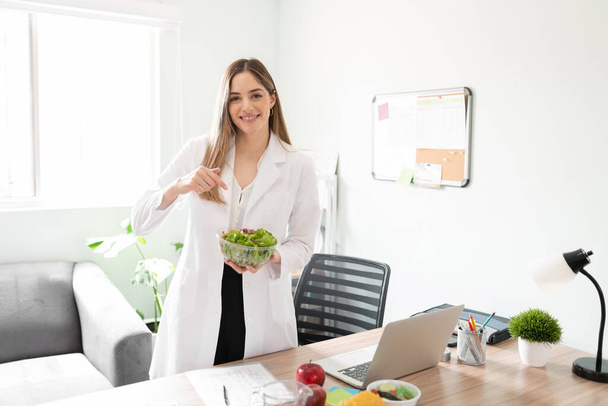 Χαριτωμένη γυναίκα διαιτολόγος δείχνοντας σε ένα μπολ σαλάτα και συνιστώντας να τρώνε περισσότερα λαχανικά και πιο υγιή - Φωτογραφία, εικόνα