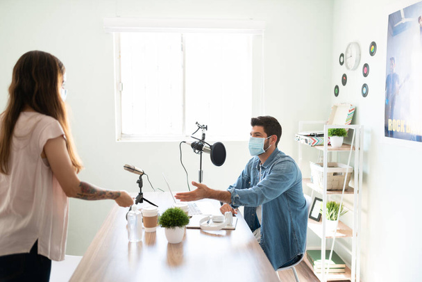 Portrait of a male radio host and announcer inviting his guest to sit down for an interview while both wearing masks during the covid19 pandemic - Photo, Image