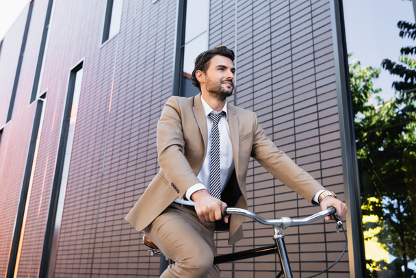 low angle view of bearded man in suit riding bicycle, smiling and looking away near building  - Фото, изображение