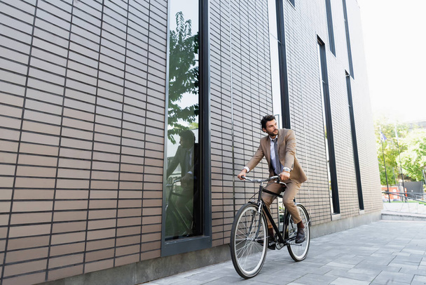 full length of businessman in suit riding bicycle and looking away near building  - Foto, afbeelding