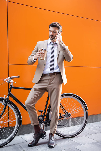 pleine longueur de l'homme d'affaires parlant sur smartphone et tenant tasse en papier près de vélo et bâtiment avec des murs orange - Photo, image