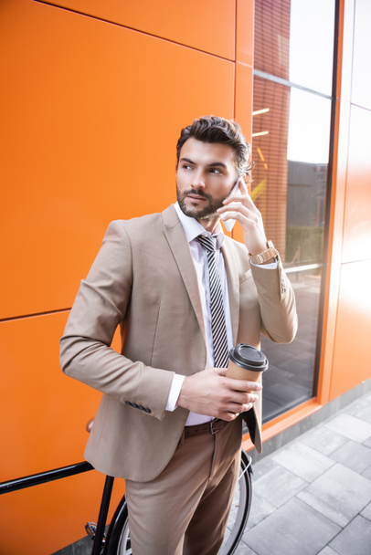 με αυτοπεποίθηση επιχειρηματίας μιλώντας στο smartphone και κρατώντας χάρτινο κύπελλο κοντά ποδήλατο και κτίριο με πορτοκαλί τοίχους - Φωτογραφία, εικόνα
