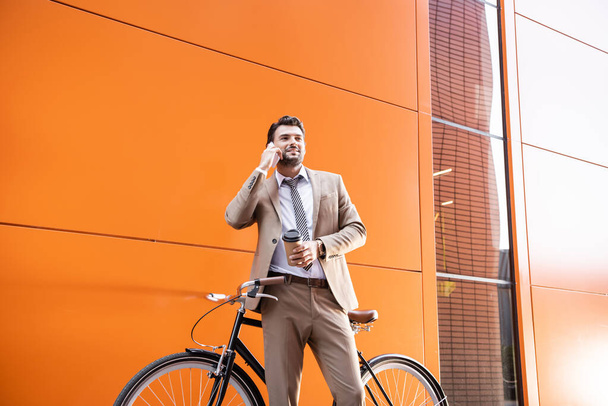 glimlachende zakenman praat op smartphone en houdt papieren beker in de buurt van fiets en gebouw met oranje muren - Foto, afbeelding