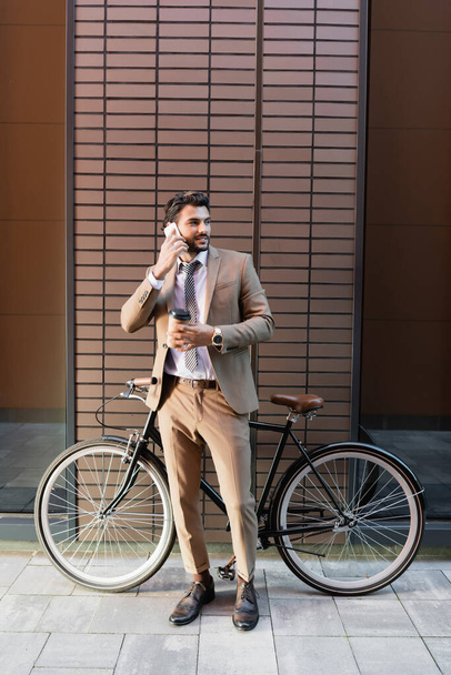 pełna długość brodatego biznesmena rozmawiającego na smartfonie i trzymającego papierową filiżankę w pobliżu roweru i budynku  - Zdjęcie, obraz