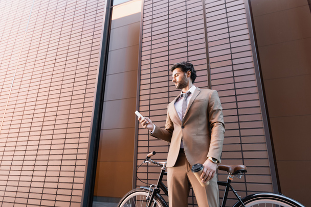 γενειοφόρος επιχειρηματίας κοιτάζοντας smartphone και κρατώντας χάρτινο κύπελλο κοντά ποδήλατο, ενώ στέκεται κοντά στο κτίριο  - Φωτογραφία, εικόνα