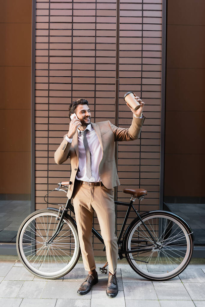 pełna długość szczęśliwy biznesmen rozmawiając na smartfonie i trzymając papierową filiżankę w pobliżu roweru i budynku  - Zdjęcie, obraz