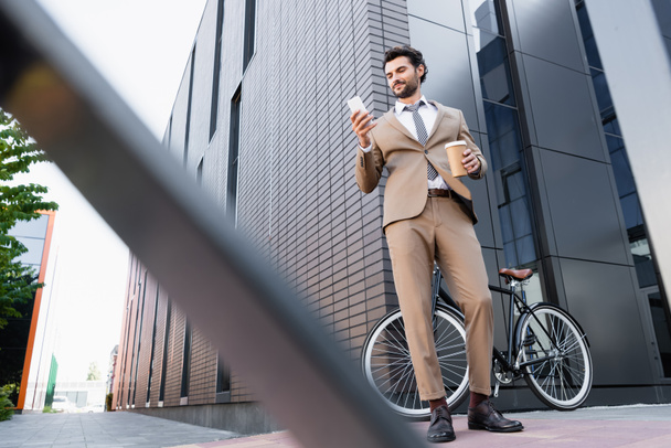 low angle view of cheerful businessman looking at smartphone and holding paper cup near bike and blurred foreground  - Фото, изображение
