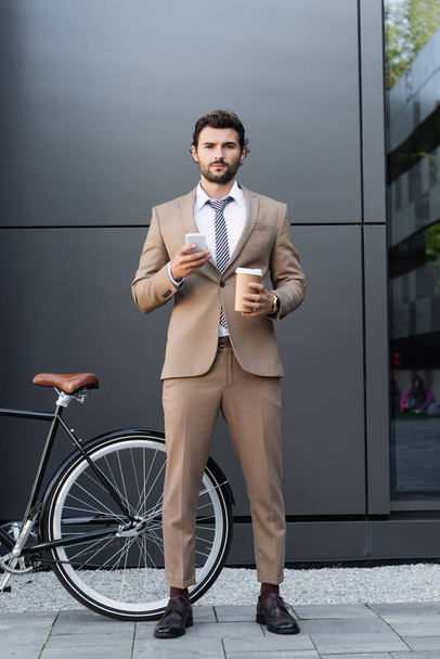 full length of bearded businessman holding coffee to go and smartphone near bicycle  - Фото, зображення