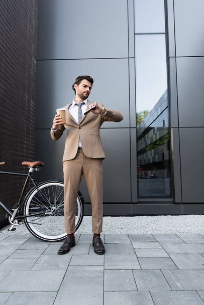 longitud completa de hombre de negocios barbudo en traje sosteniendo café para ir y teléfono inteligente mientras mira el reloj cerca de la bicicleta  - Foto, Imagen