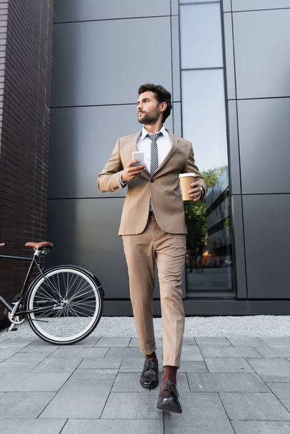 full length of businessman in suit holding coffee to go and smartphone while walking near bicycle  - Zdjęcie, obraz