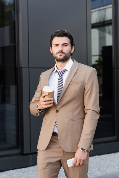 bearded businessman in formal wear holding coffee to go and smartphone near modern building - Foto, Bild