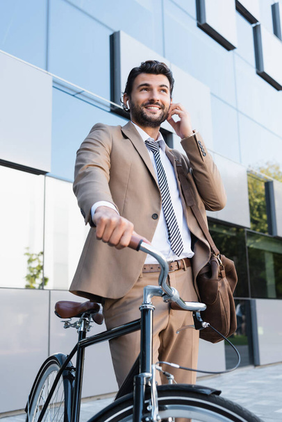low angle view of smiling businessman in wireless earphones while standing with bag near bike and building - Photo, Image