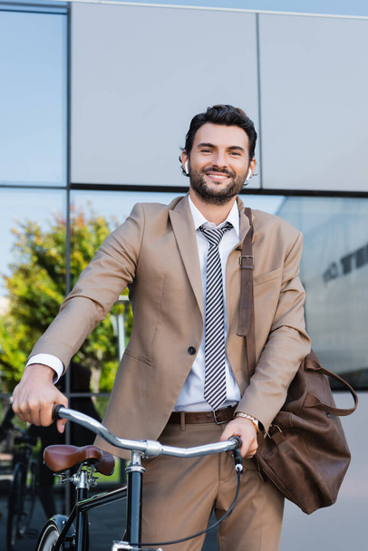 χαμογελαστός επιχειρηματίας σε ασύρματα ακουστικά ενώ στέκεται με τσάντα κοντά σε ποδήλατο και κτίριο - Φωτογραφία, εικόνα