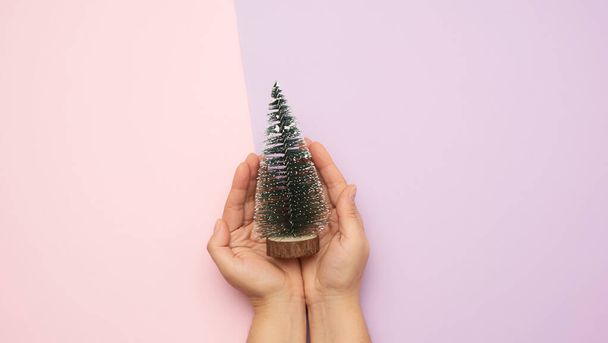 manos sosteniendo la decoración de Navidad árbol verde en la superficie de color, minimalismo, vista superior - Foto, Imagen