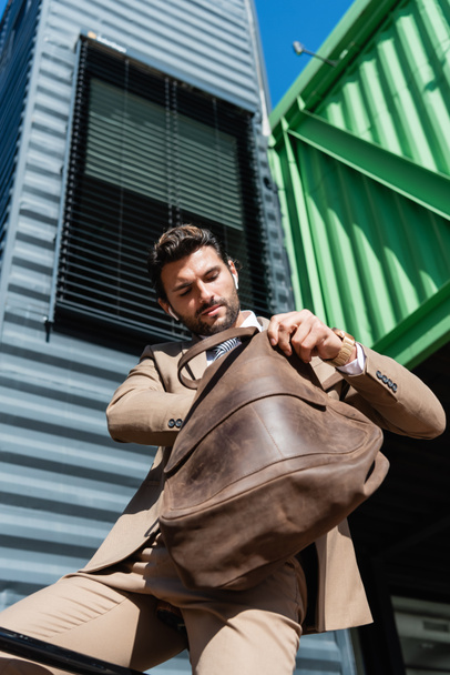 low angle view of bearded businessman in wireless earphones holding leather bag  - Photo, Image