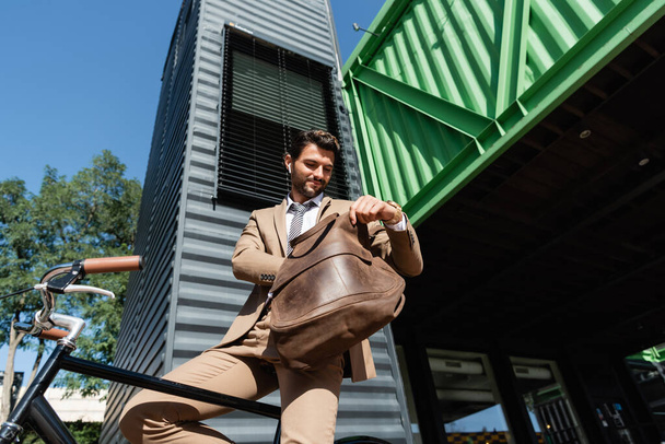 low angle view of cheerful businessman in wireless earphones holding leather bag near bike - Foto, afbeelding