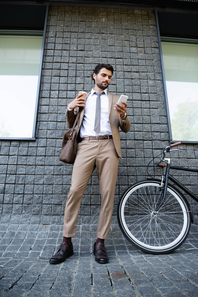 full length of businessman in wireless earphones and suit holding paper cup and smartphone near building  - Foto, imagen