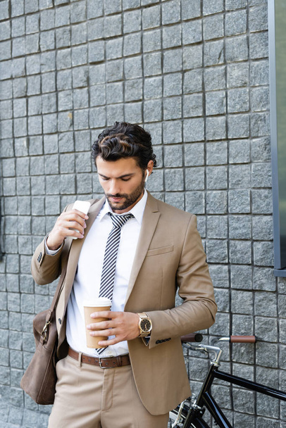 uomo d'affari in auricolari wireless e abbigliamento formale che tiene il caffè per andare e smartphone vicino edificio  - Foto, immagini