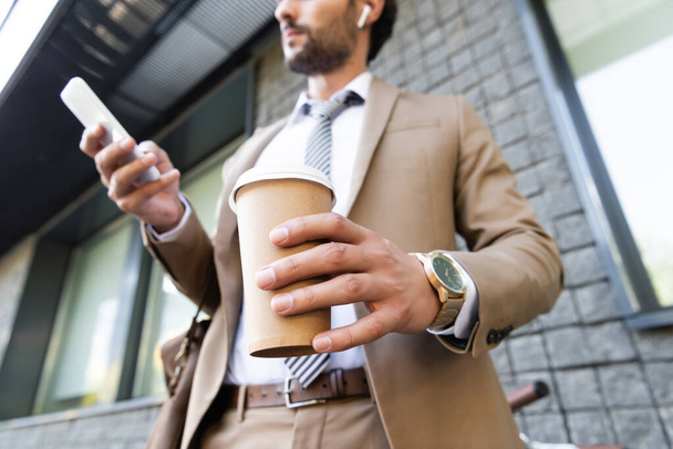 cropped view of businessman in wireless earphones and suit holding coffee to go and smartphone near building  - Fotografie, Obrázek
