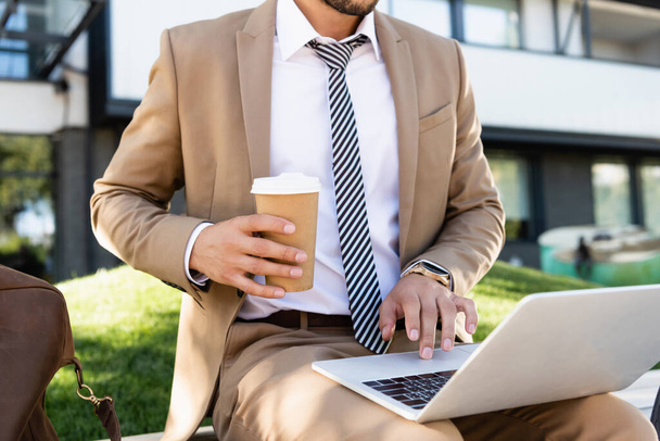 vista recortada del hombre de negocios utilizando el ordenador portátil y la celebración de la taza de papel - Foto, Imagen