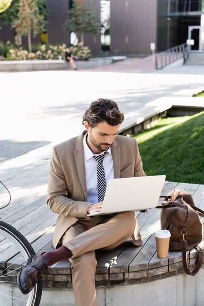 businessman in suit using laptop while sitting on bench with paper cup and bag - Foto, immagini