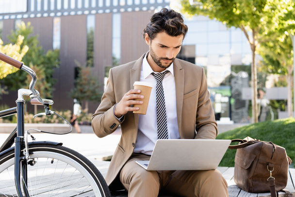 γενειοφόρος επιχειρηματίας με κοστούμι χρησιμοποιώντας φορητό υπολογιστή και κρατώντας χάρτινο κύπελλο, ενώ κάθεται στον πάγκο κοντά στο ποδήλατο - Φωτογραφία, εικόνα