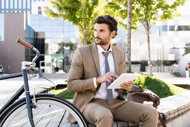 üzletember öltönyben digitális tabletta, miközben ül padon közel kerékpár  - Fotó, kép