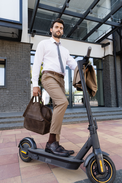 full length of businessman in formal wear standing near e-scooter with leather bag  - Foto, afbeelding