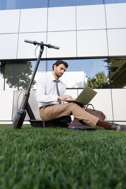 businessman in formal wear sitting with laptop near e-scooter on grass - Zdjęcie, obraz