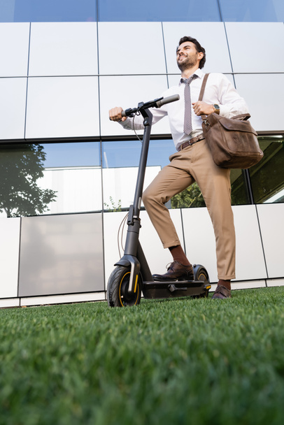 полная длина счастливого бизнесмена в формальной одежде стоя рядом с электрическим скутером на траве  - Фото, изображение