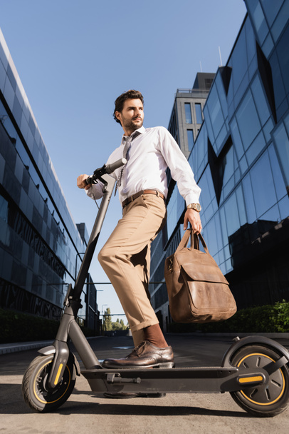 low angle view of businessman in formal wear holding leather bag and standing near electric scooter  - Foto, Imagen