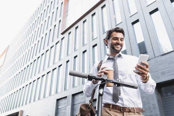 low angle view of happy businessman in formal wear holding paper cup and using smartphone near e-scooter  - Фото, изображение