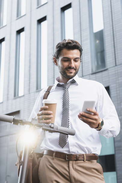 Улыбающийся бизнесмен в формальной одежде держит бумажную чашку и использует смартфон возле электронного скутера  - Фото, изображение